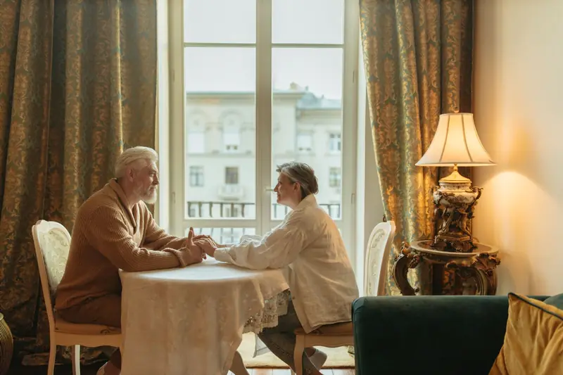 An older couple hold hands across a small table by a sunlit window
