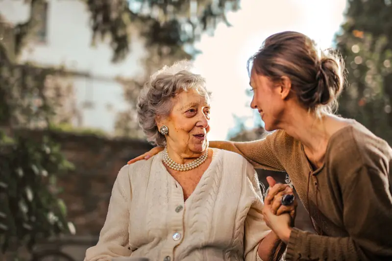 An adult daughter warmly greets her smiling elderly mother in a sunny garden