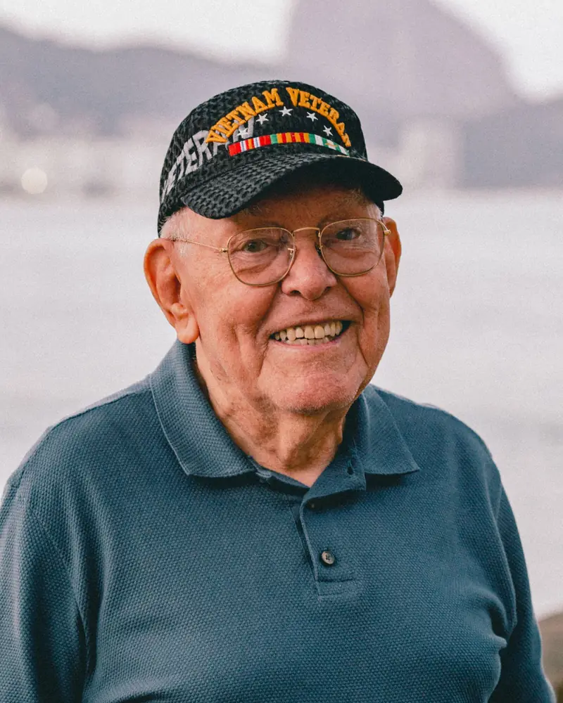 A smiling older man wearing a Vietnam veteran cap by the ocean